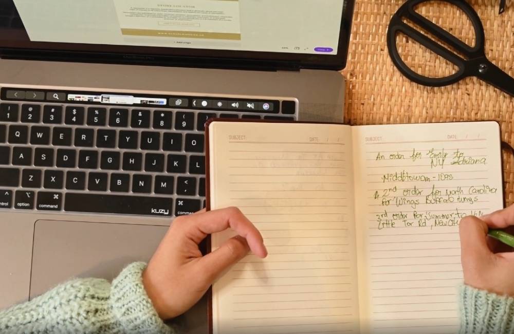 ノートパソコンの前で、一人の人物がノートに手書きでメモやレポートを書き込んでいる様子。