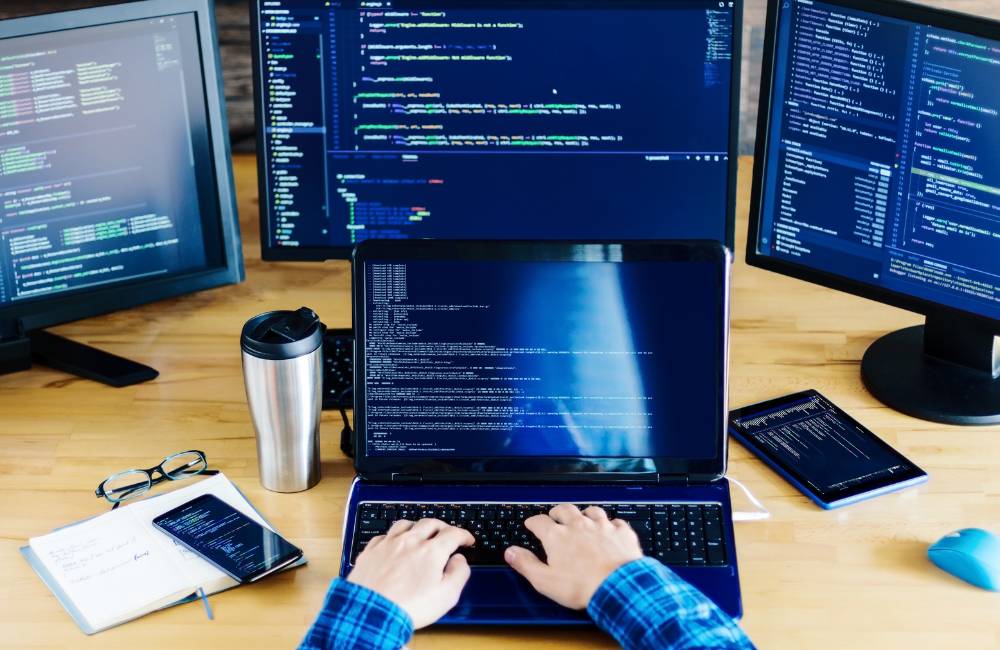 複数の外部モニターにコードが表示された、プログラミングや開発作業のためのデスク環境。手前では、チェック柄のシャツを着た人物がノートパソコンを操作している。