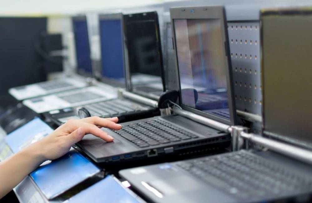 中古ノートPCが多数並ぶ陳列棚。ユーザーがPCのキーボードに手を添えて状態を確認しており、在庫入れ替え時期の選定の様子。