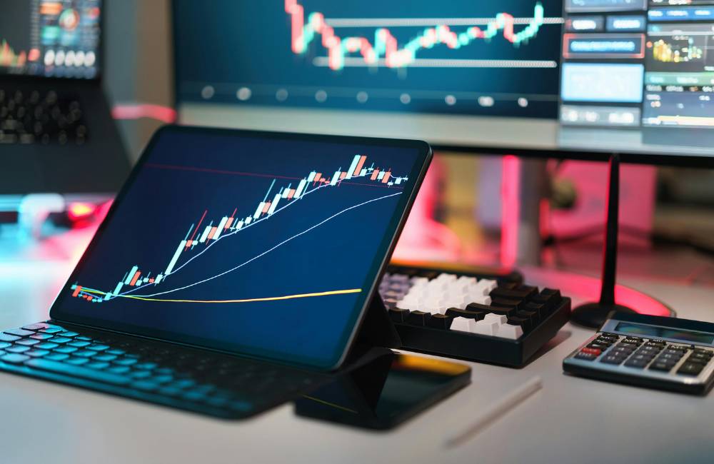 タブレットとPCの画面に価格変動を示すグラフ（ローソク足チャート）が表示されたデスク。中古PC市場の価格推移や変動傾向を分析するイメージ。