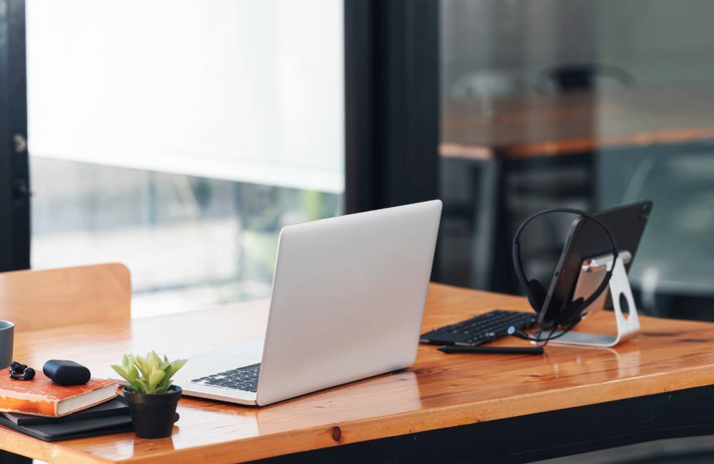 窓際にある木製のデスクに置かれたシルバーのノートPC。観葉植物やヘッドフォンがあり、安心感と快適さを備えた中古PCの使用環境。