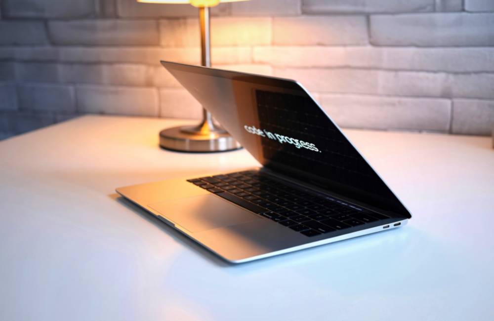 テーブルランプに照らされたモダンなノートパソコンが開かれており、画面には「code in progress.」と表示されているスタイリッシュな様子