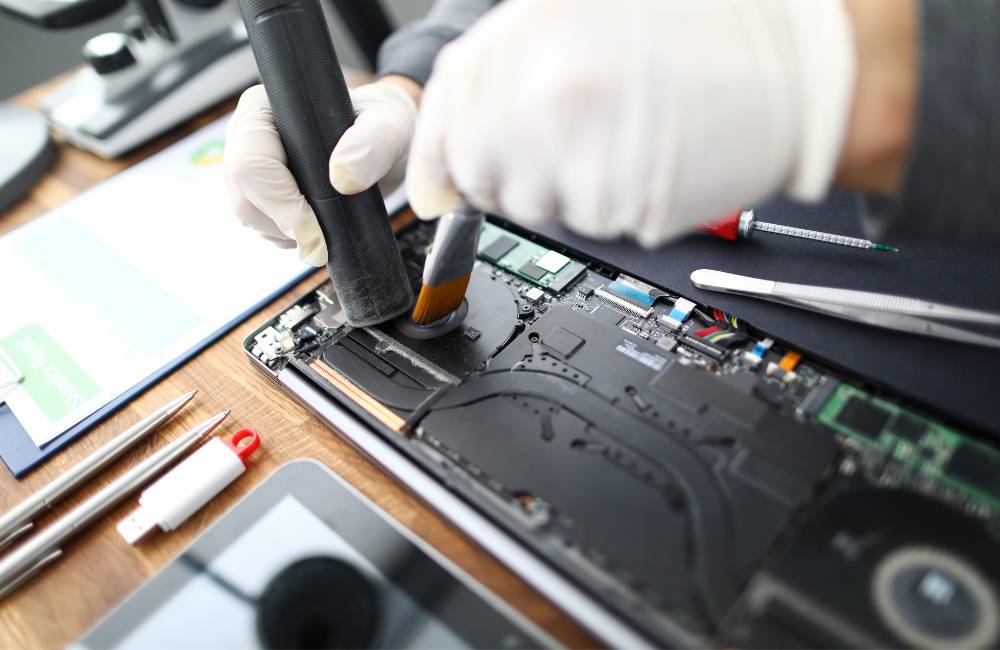 白い手袋をした技術者が、ノートパソコンの内部基板を吸引ノズルとブラシを使って丁寧にクリーニングしている様子