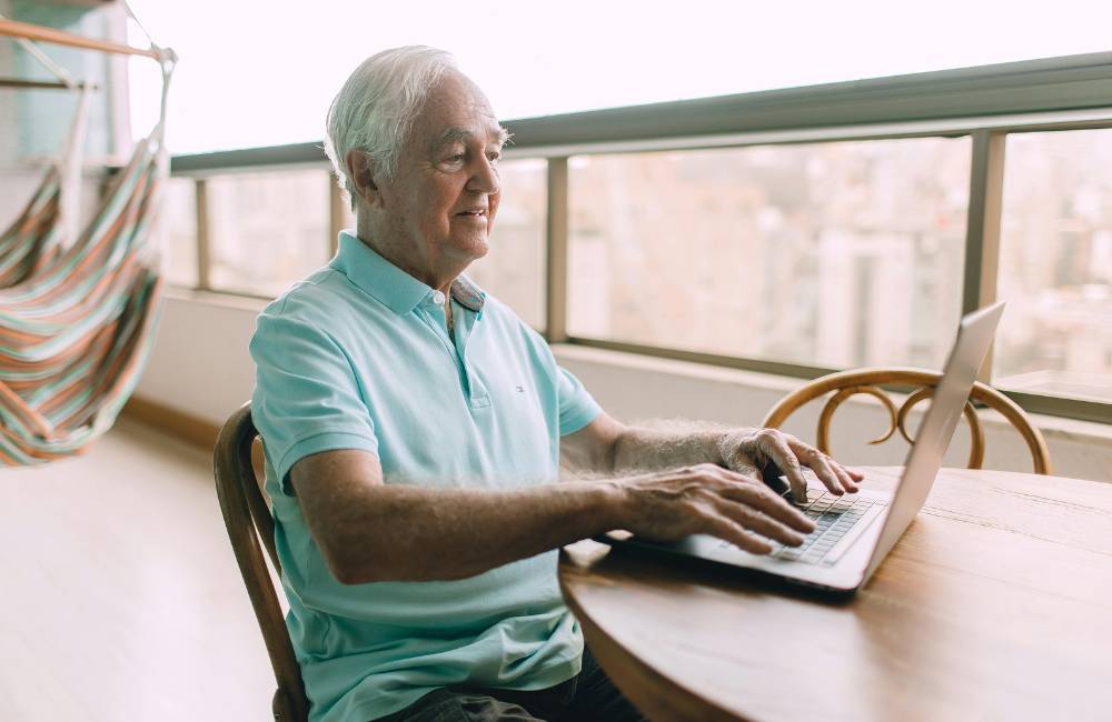 バルコニーのテーブルで、笑顔の年配の男性がノートパソコンを快適に操作している様子。中古PC選びに成功した理想のイメージ。