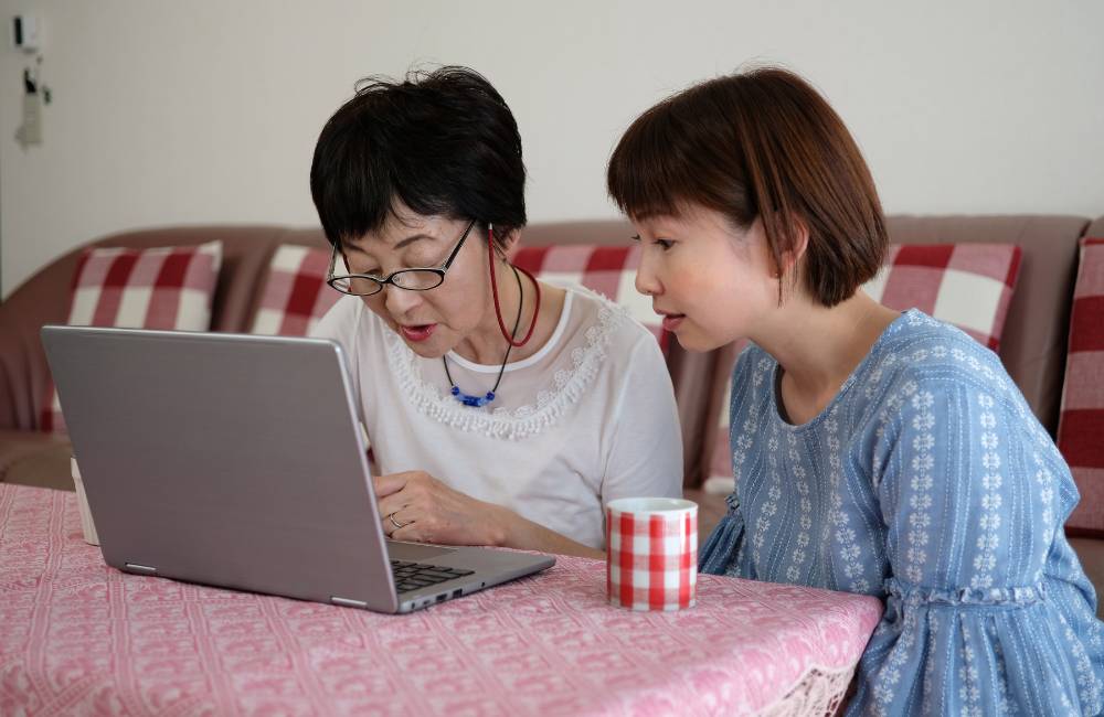 リビングで、若い女性が年配の女性にノートパソコンの操作を熱心に教えている様子。家族によるデジタルサポートの重要性。