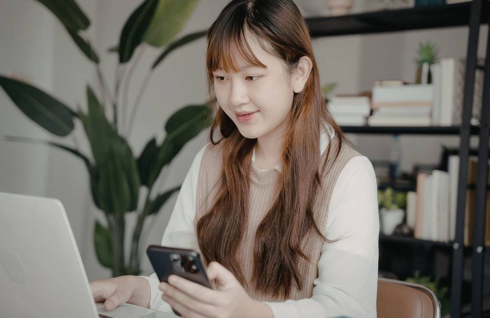 ノートパソコンの前でスマートフォンを操作しながら、穏やかに微笑む女性の姿。初心者でも扱いやすいPCモデルのイメージ。