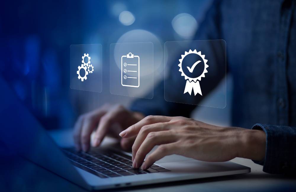 ノートパソコンを操作する手の画像の上に、歯車（プロセス）、チェックリスト、品質保証の認定証アイコンがデジタルオーバーレイで表示されている。
