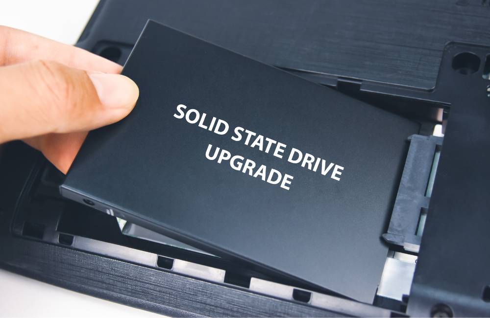 ノートパソコンの裏蓋を開け、「SOLID STATE DRIVE UPGRADE」と書かれたSSD（ソリッドステートドライブ）を換装している手のクローズアップ。