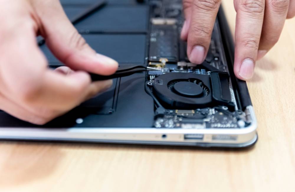 ピンセットを使い、分解されたノートパソコン内部の冷却ファンや精密基盤に手を加えている修理作業のクローズアップ。