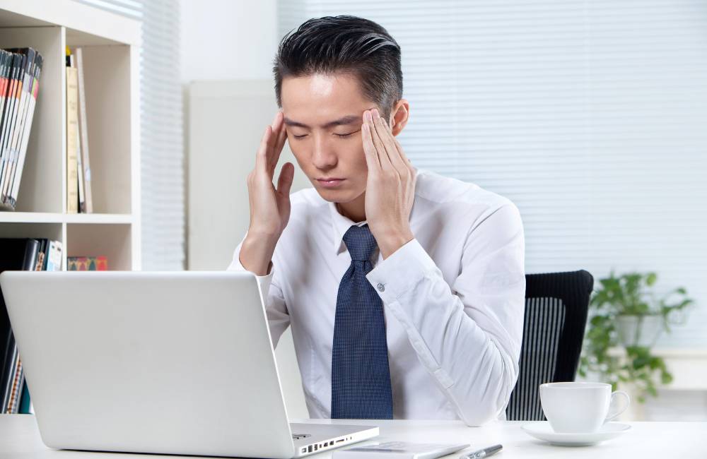 職場でノートパソコンの前に座り、ストレスや頭痛でこめかみを押さえているビジネスマン。PCの不調によるイライラを表現。