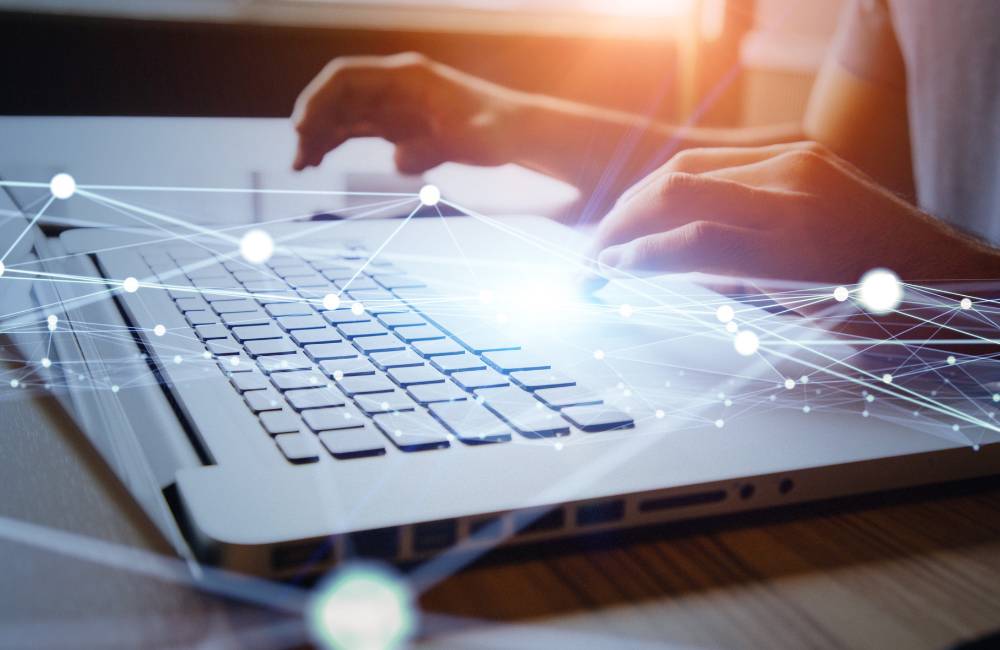 ノートパソコンのキーボードを操作する手に、ネットワーク接続を思わせる光のグラフィックが重ねられた画像。ネットワーク設定やOSの設定を確認する様子。