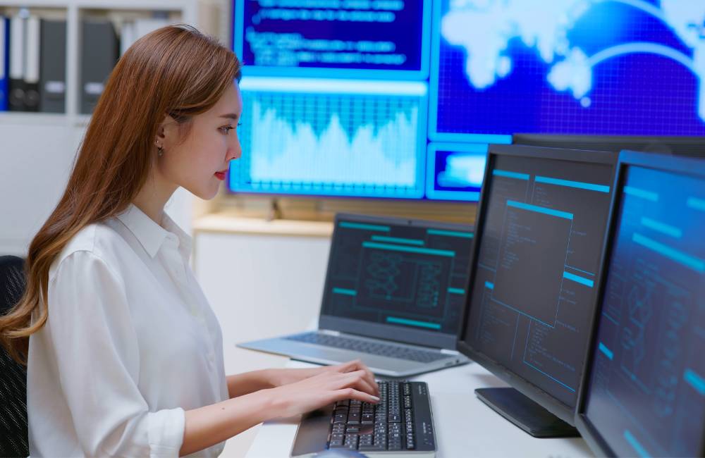 複数の監視モニターとキーボードを操作する女性のセキュリティ担当者。システムの監視と防御設定の管理を行っています。