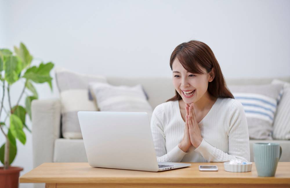 リビングでノートPCを見つめ、笑顔で手を合わせる女性。セキュリティ対策後の安心感と快適なPC利用を象徴しています。