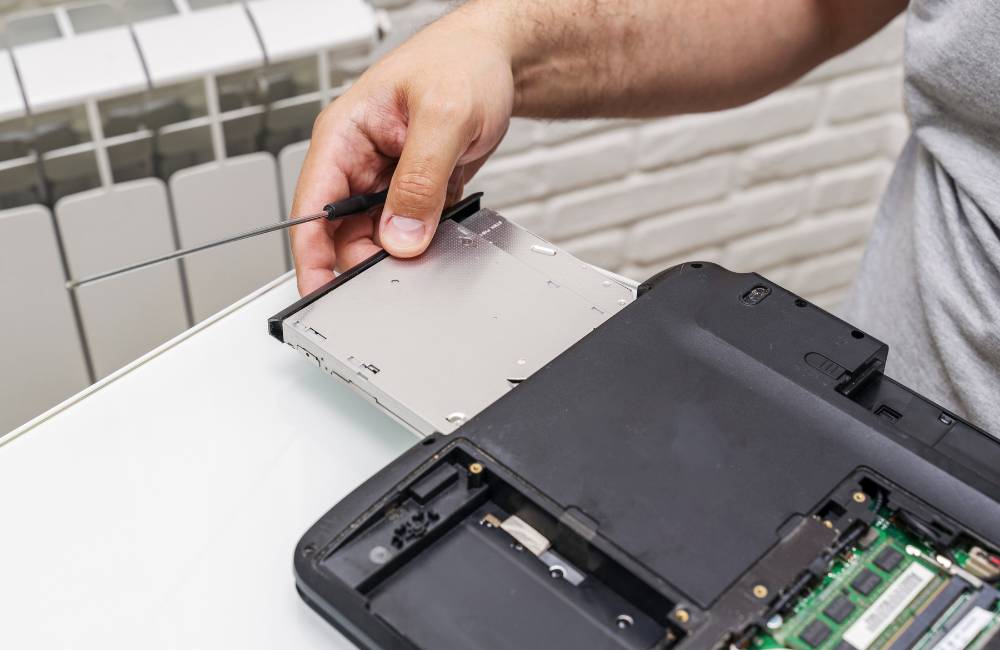 ノートパソコンの裏蓋を開け、手でHDDまたはSSDドライブベイ（ストレージ）を取り外しているクローズアップ。PCの内部パーツを確認している様子。