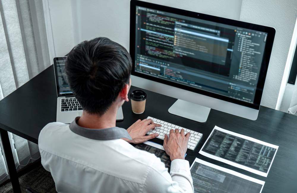 男性がデスクで複数のモニターを使い、専門的なプログラムコードやシステム設定を真剣な表情で確認・操作している様子。