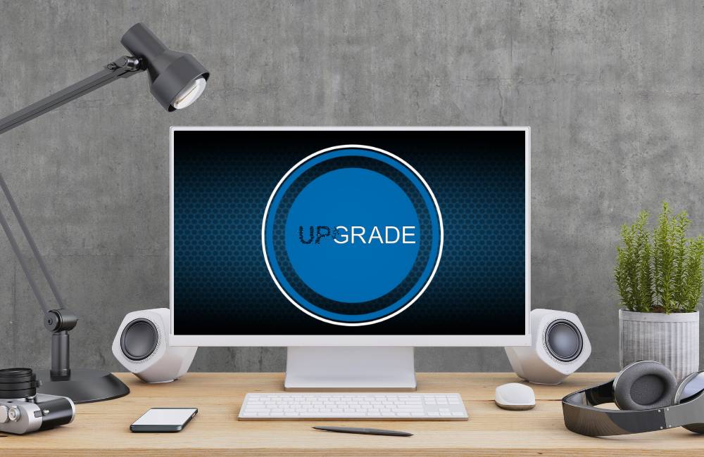 中央に青い丸と「UPGRADE」の文字が表示されたデスクトップPCのモニター。モダンなデスクに設置され、アップグレードの準備や検討を表現。