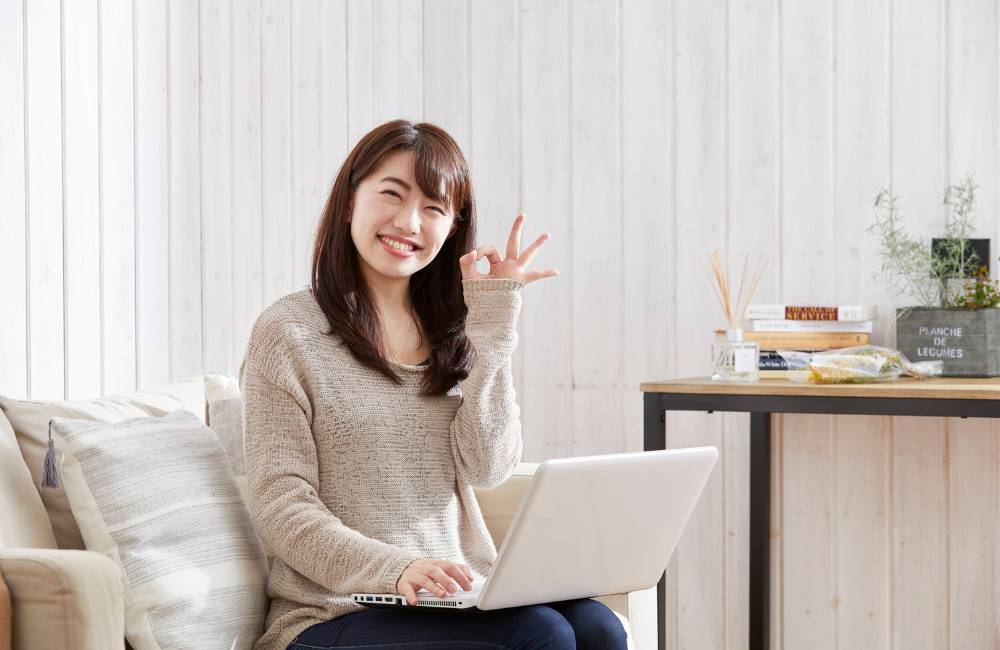 女性がソファに座り、白いノートPCを膝に置いて、笑顔でカメラに向かってOKサイン（サムズアップ）をしている。