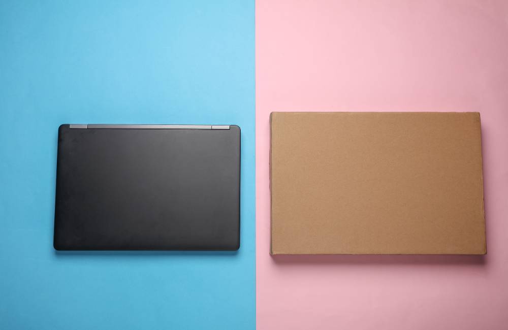 青とピンクの鮮やかな背景の上に、黒いノートパソコン本体と、その横に茶色の段ボール箱が置かれている様子。購入品と梱包。