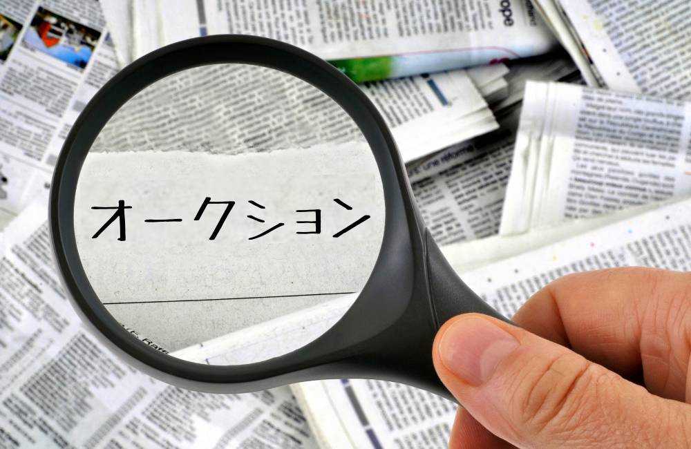 雑然とした新聞記事の上に置かれた虫眼鏡で、紙片に書かれた「オークション」の文字を拡大している様子。