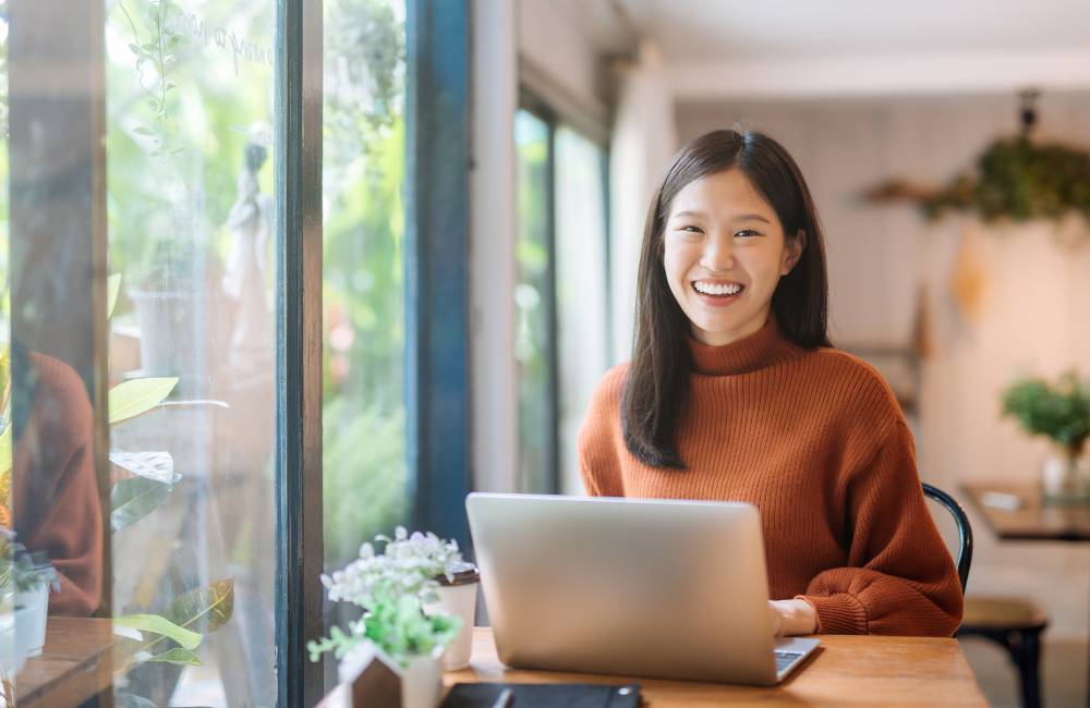 カフェのような場所で、ノートパソコンの前で笑顔でカメラを見ている女性。購入後の満足感やリユースの明るいイメージ。