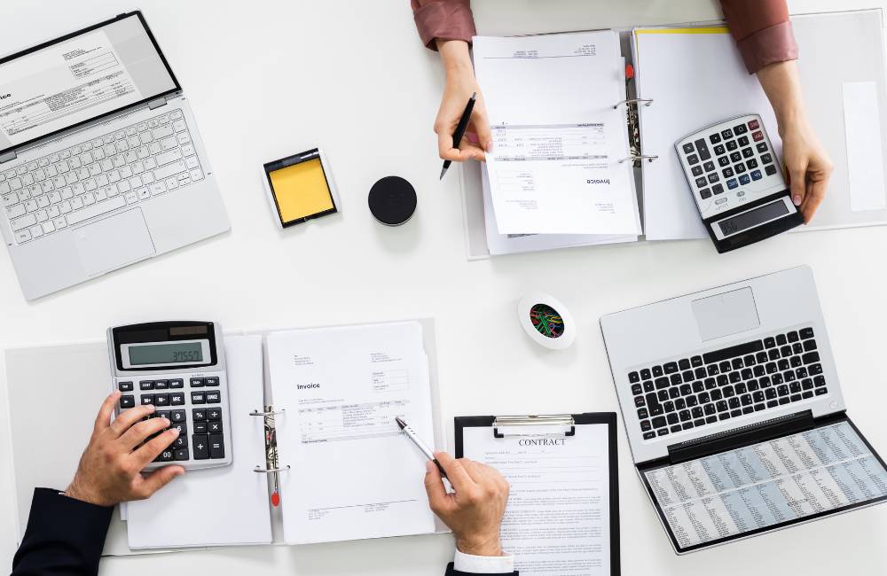 白いデスクの上で、2台のノートパソコン、2台の電卓、契約書、請求書などの書類を整理・操作している2人の人物を真上から捉えた俯瞰写真