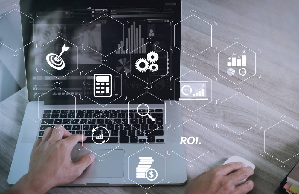 ノートパソコンを操作する手元に、ターゲット、歯車、計算機、ROI（投資利益率）などのビジネスアイコンがデジタル合成されたイメージ画像