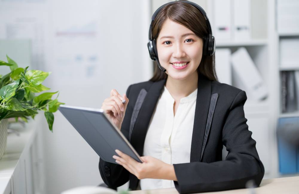 オフィスでヘッドセットを装着し、タブレットとペンを手に持ちながら微笑む、カスタマーサポート担当の女性