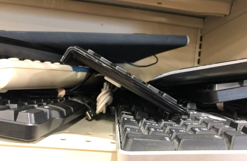 中古ショップの棚に雑然と積み上げられた、清掃・整備前のキーボードやPCパーツなどのジャンク品。