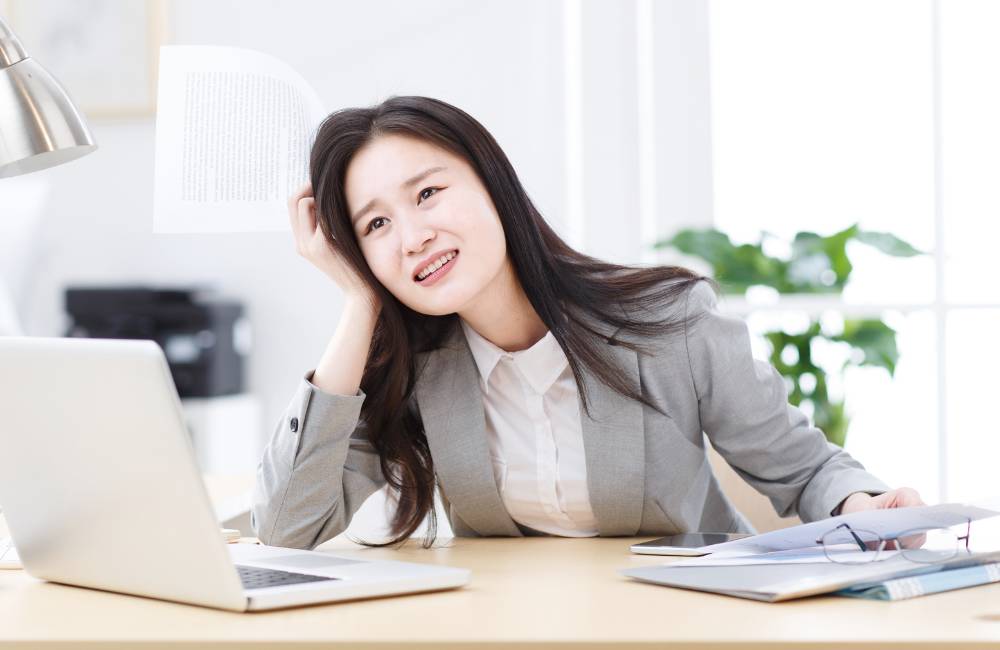 パソコンを前に書類を手に持ちながら、保証書を失くして困り果てた表情で頭を抱えるビジネスウェアの女性