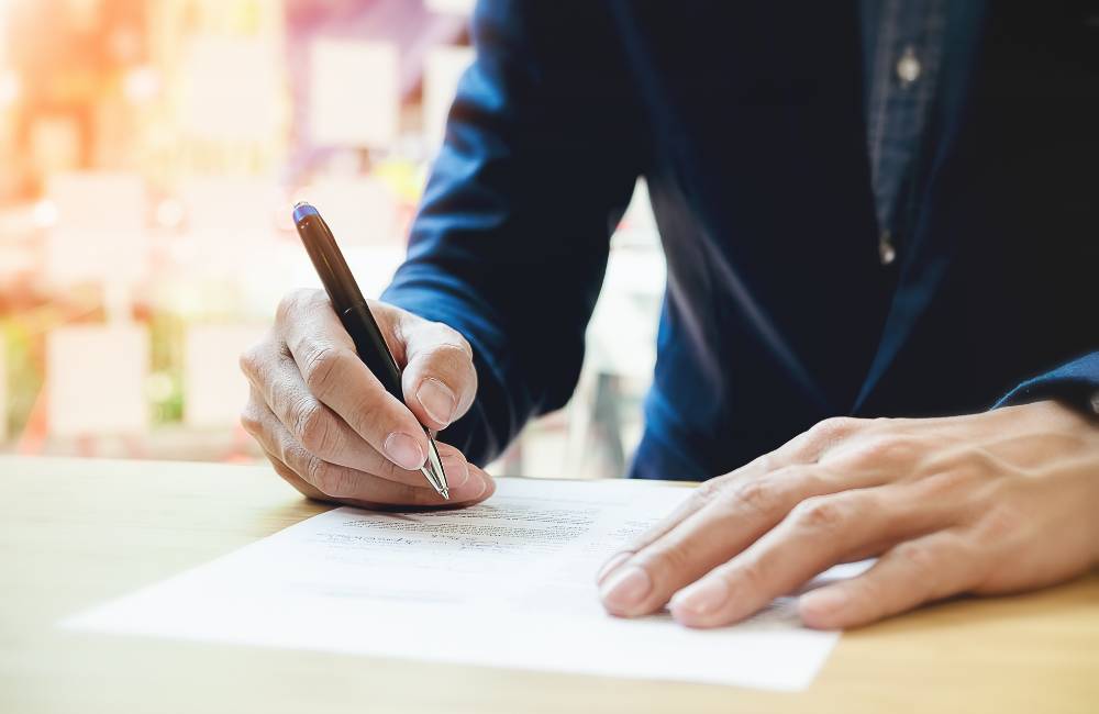 スーツを着た人物が、机の上の書類にボールペンで署名（サイン）をしている手元のクローズアップ