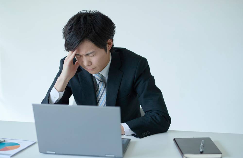 ノートPCの前で頭を抱えて不安そうな表情をしているスーツ姿の男性