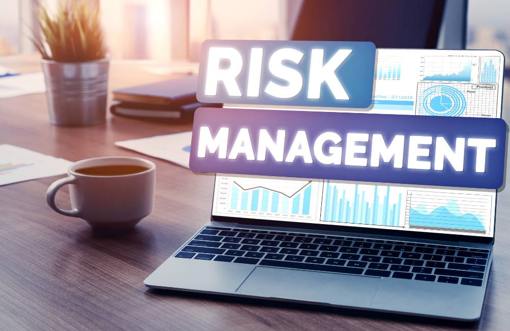 デスク上のノートパソコンの画面に「RISK MANAGEMENT（リスク管理）」の文字と、分析用の統計グラフやチャートが表示されている様子