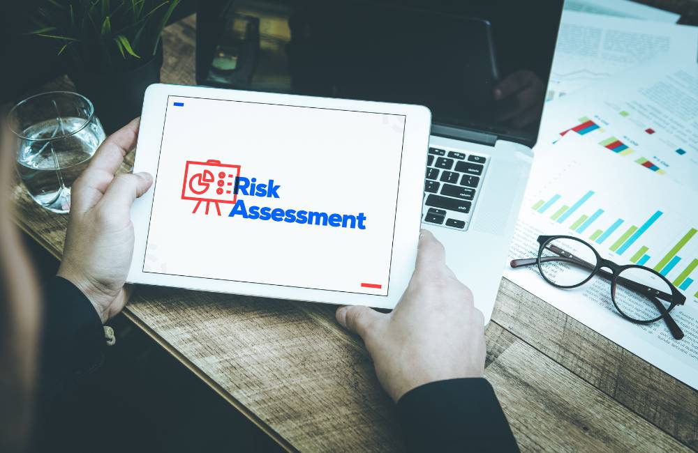 木目調のデスクで、タブレットに表示された「Risk Assessment（リスク評価）」のロゴとグラフを両手で持って確認している様子