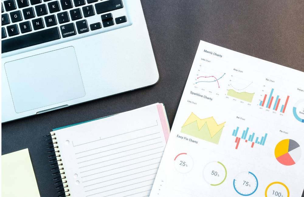 黒いデスクの上に置かれたノートパソコンの一部と、複数の円グラフや折れ線グラフが印刷された分析レポート、および罫線ノートの俯瞰図