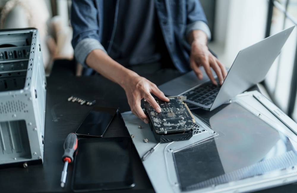 整備環境でノートパソコンを操作しながら、もう一方の手でパソコンの内部パーツ（グラフィックボード）を素手で持ち上げている様子