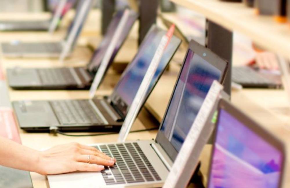 パソコンショップの店頭にさまざまな種類のノートパソコンが並んで陳列されており、購入者が手前のパソコンのキーボードに触れて確認している様子