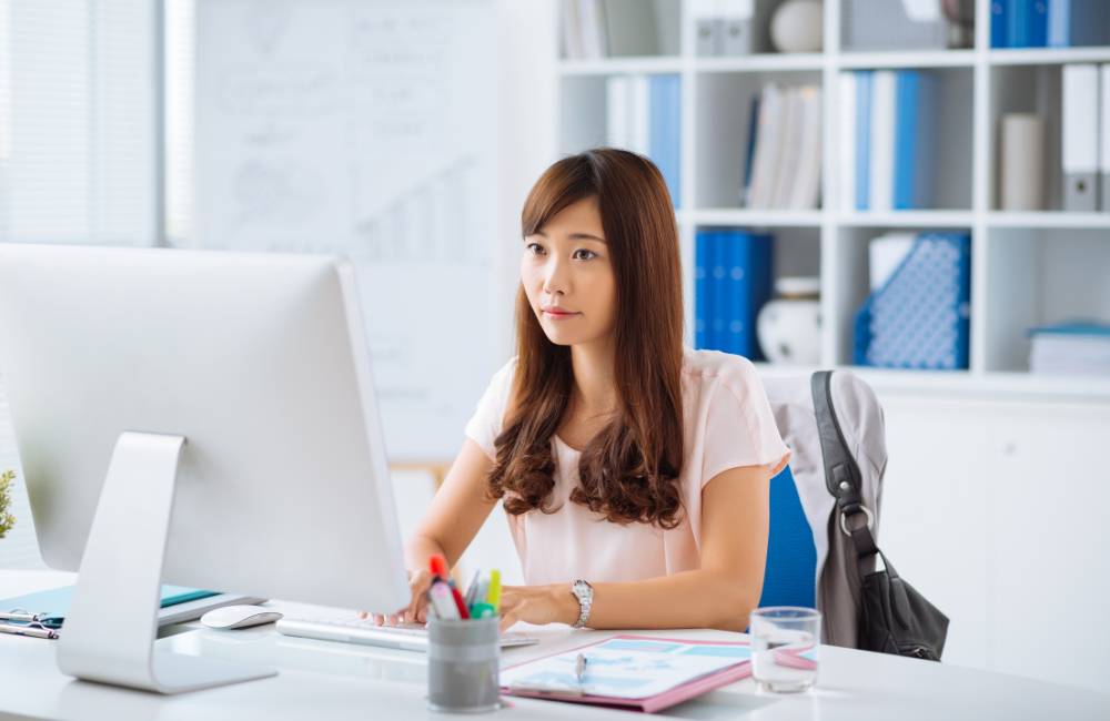 オフィスで、女性が集中してデスクトップパソコンの画面を見ながらキーボードを操作している。