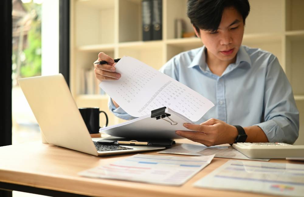 明るい室内でノートパソコンを操作し、書類を確認しながら作業する男性の様子