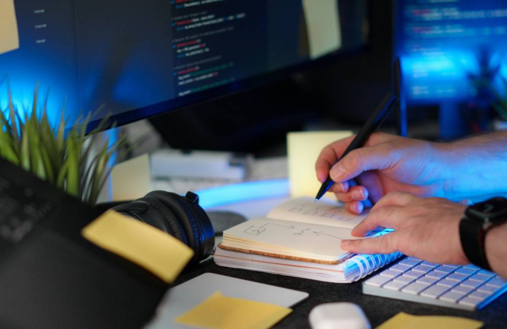 数のモニターが並ぶデスクで、ノートを開きながらペンで図解やアイデアを書き留めているクリエイティブな作業風景