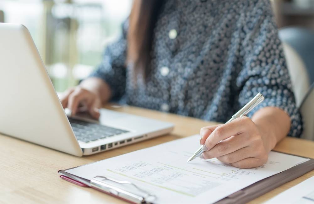 ノートパソコンのキーボードと、クリップボードに固定された書類にペンで書き込みをする手元のアップ