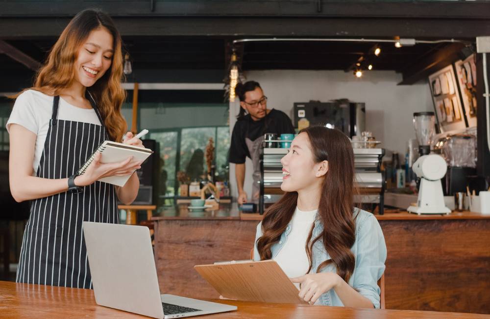 カフェのような空間で、店員と客がノートPCの画面を見ながら話している様子。中古PCショップでの相談や購入・導入方法。