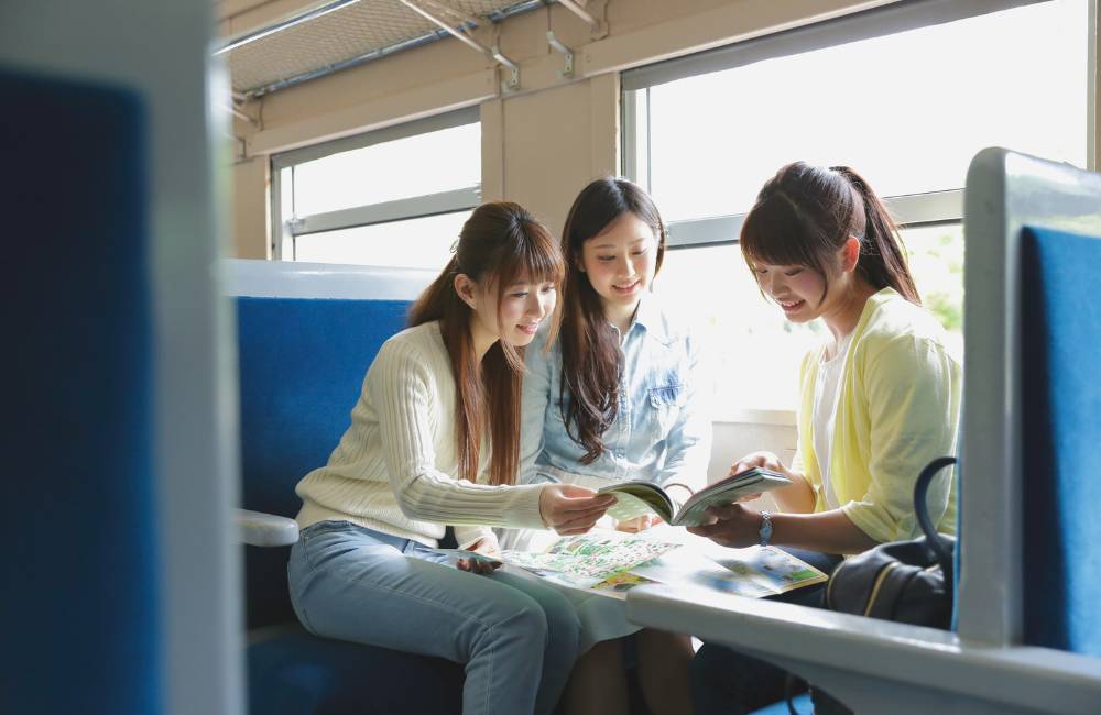 走行中の列車の座席で、旅のガイドブックを広げて楽しそうに話す2人の若い女性。旅の思い出をVlog（ビデオブログ）にするために動画編集を始めたいアクティブな初心者のイメージ。