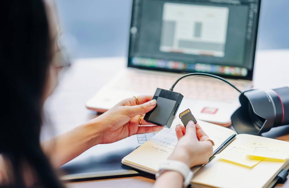 デスクの上でSDカードを手に持ち、ノートにペンで計画を書き込んでいる女性の手元。初心者が素材管理や編集のコツを学ぶ準備段階を表現。