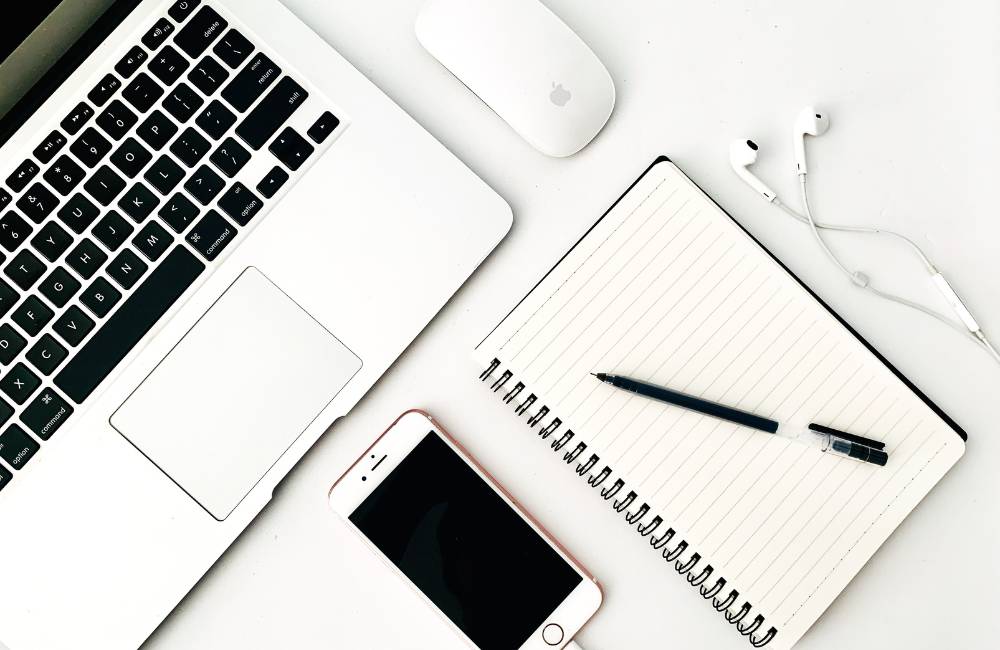 ノートパソコン、スマートフォン、イヤホン、ノートとペンが整然と並んだデスク上の写真