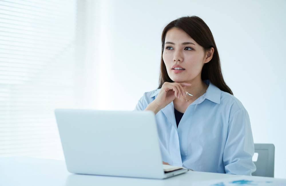 ノートパソコンを前に、顎に手を当てて考え込む女性の表情。中古かリースかの選択に悩む比較・検討のイメージ。