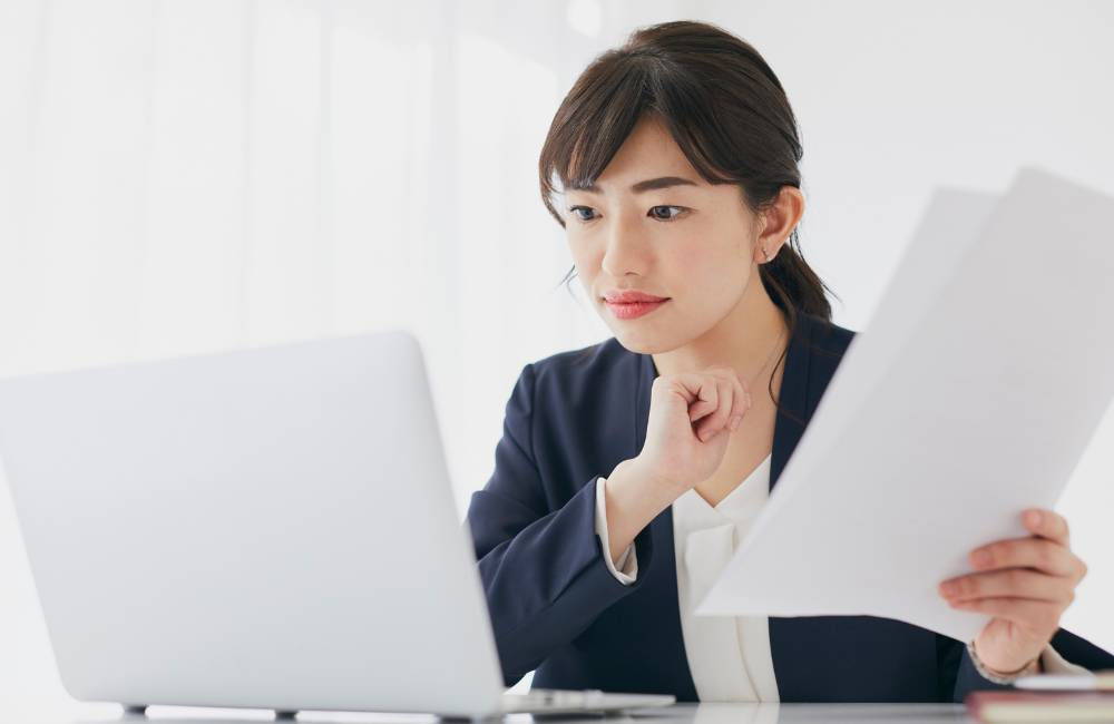 ノートPCを見ながら書類を手に持ち真剣な表情で考えるスーツ姿の女性