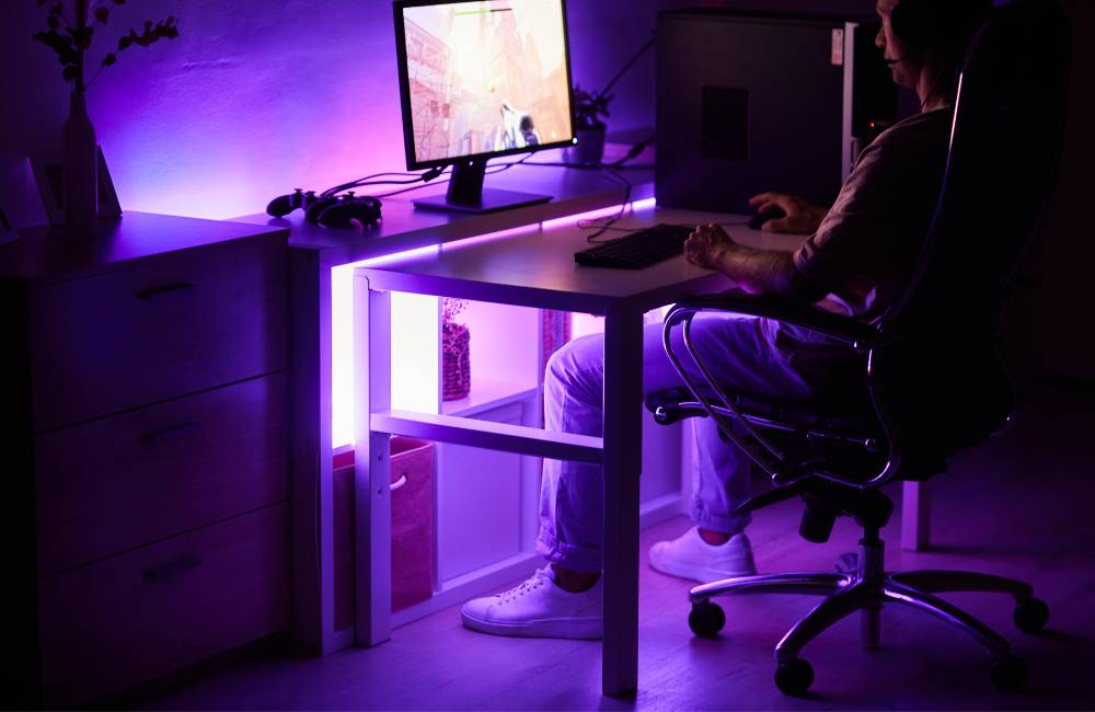 紫色の間接照明に包まれたゲーミングルームで、PCに向かって座る男性の背後からのカット。モニター、デスク、チェアが揃った中古PC予算別構成の完成イメージ。