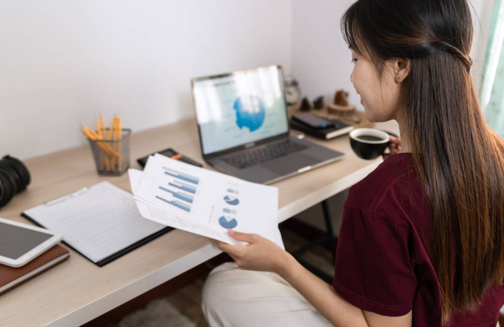 窓から光が差し込むデスクで、資料を手に取りながらコーヒーを飲む女性。背後から見た、穏やかな作業風景。