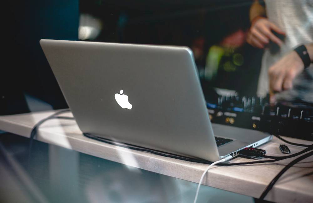 薄暗い部屋で、テーブルに置かれたMacBookのリンゴマークが光っている背面。DJミキサーのような機材が背景にぼやけて見え、夜間のクリエイティブ作業やエンターテイメント利用を連想させる。