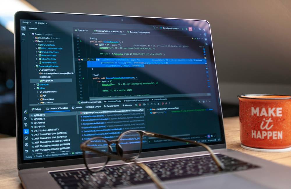 ノートパソコンの横に、「MAKE it HAPPEN」と書かれたオレンジ色のマグカップが置かれているクローズアップ写真。背景にはプログラミングコードが表示されたモニターがぼやけて写っており、目標達成へのモチベーションを感じさせる。